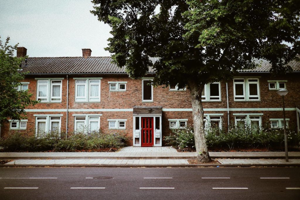 A residential brick building with a bold red front door and white-framed windows under a large green tree.